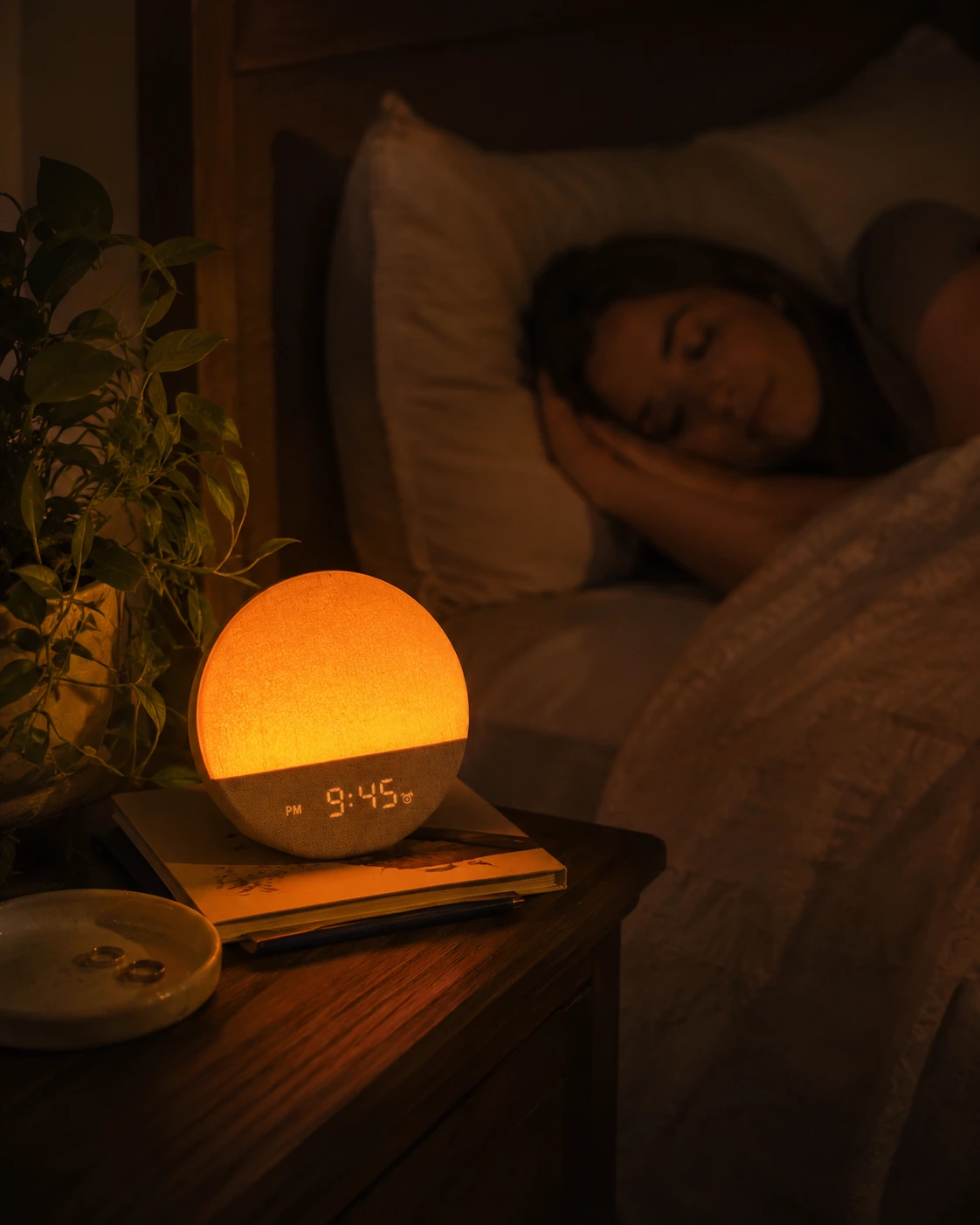 Dreamegg Sunrise 1 Wake Up Alarm Clock casting a warm orange glow on a bedside table while a person sleeps. Dreamegg Sunrise 1 Wake Up Alarm Clock casting a warm orange glow on a bedside table while a person sleeps.